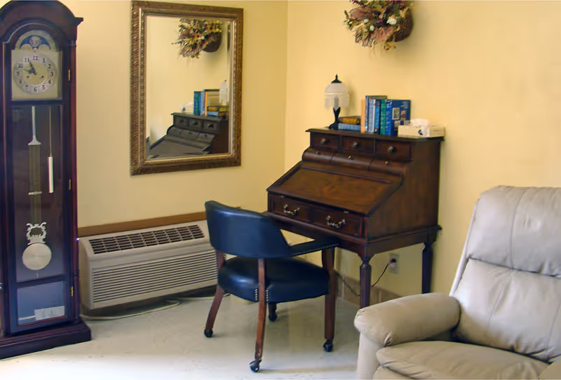 A cozy corner of a room featuring a tall grandfather clock, a wooden roll-top desk with books and a lamp on top, a black chair with wheels, a beige recliner chair, a wall-mounted mirror, and a decorative floral arrangement on the wall.