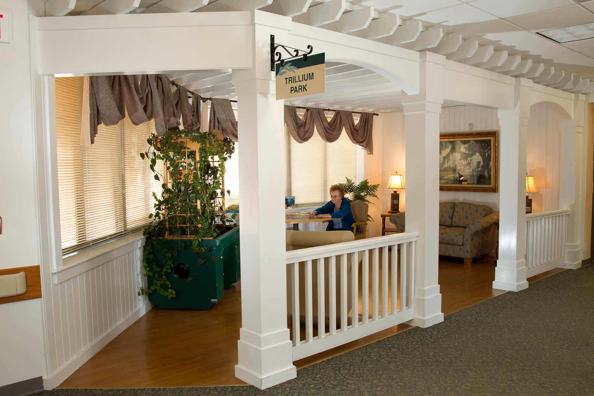 Interior common area with white columns, railing, plant, and a resident seated at a table.
