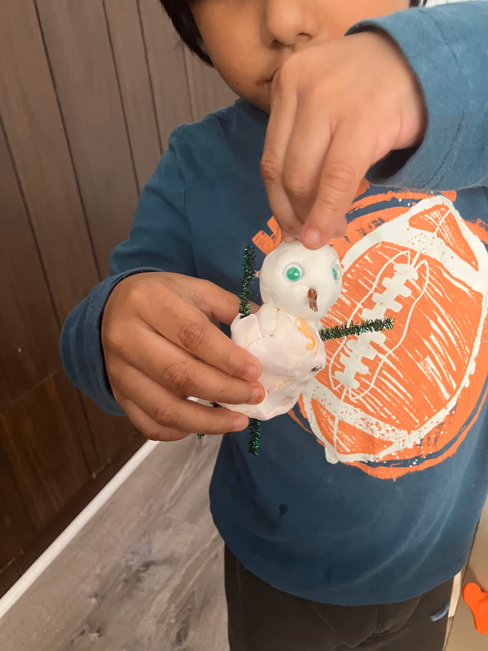 Child holds a small snowman craft with pipe-cleaner arms indoors.