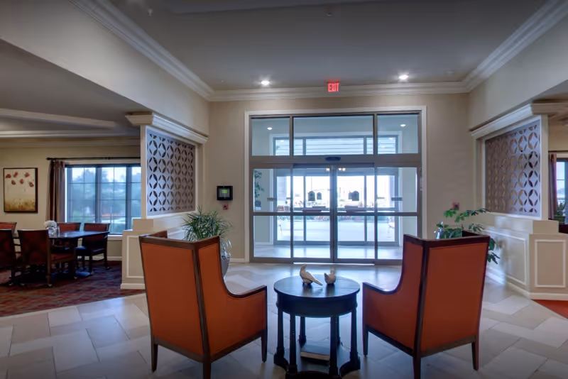 Lobby seating area with two orange chairs facing a small round table and glass entry doors.
