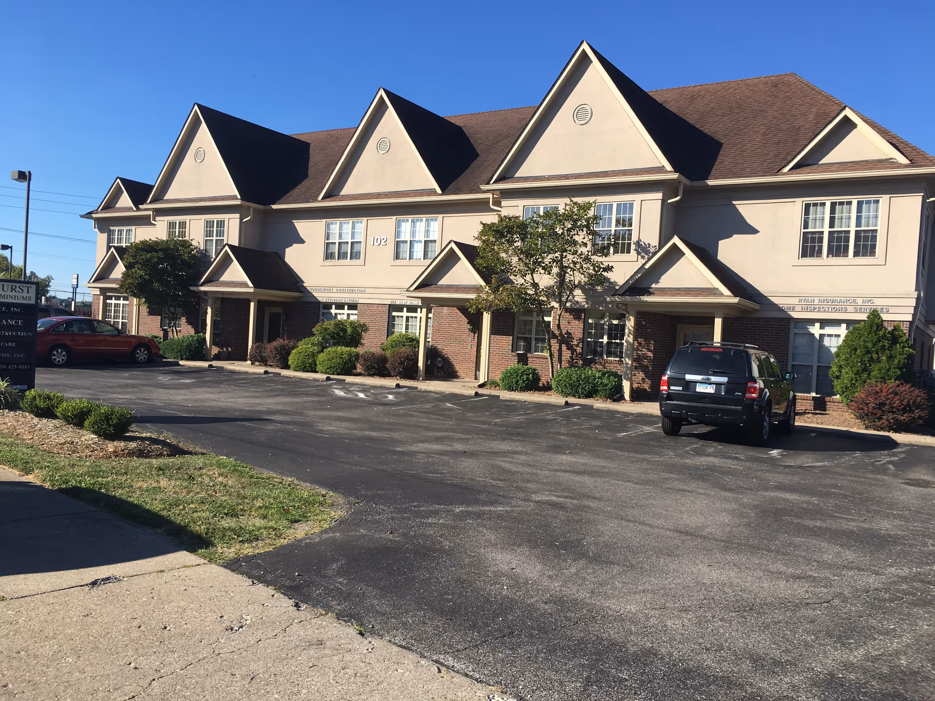 Two-story beige senior care building with pointed roofs, parking lot, and shrubs.