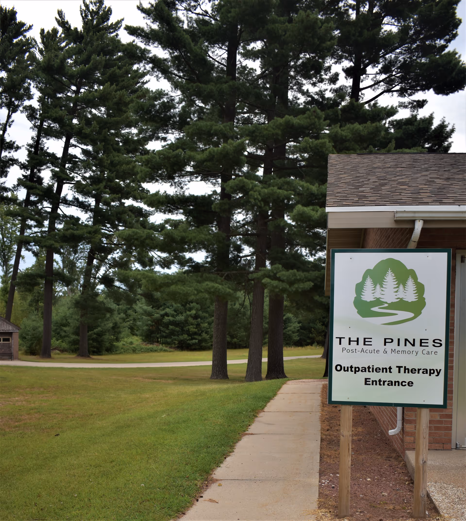 Pathway and lawn lined with tall pine trees beside a building and a sign reading 'The Pines Post-Acute & Memory Care Outpatient Therapy Entrance'.