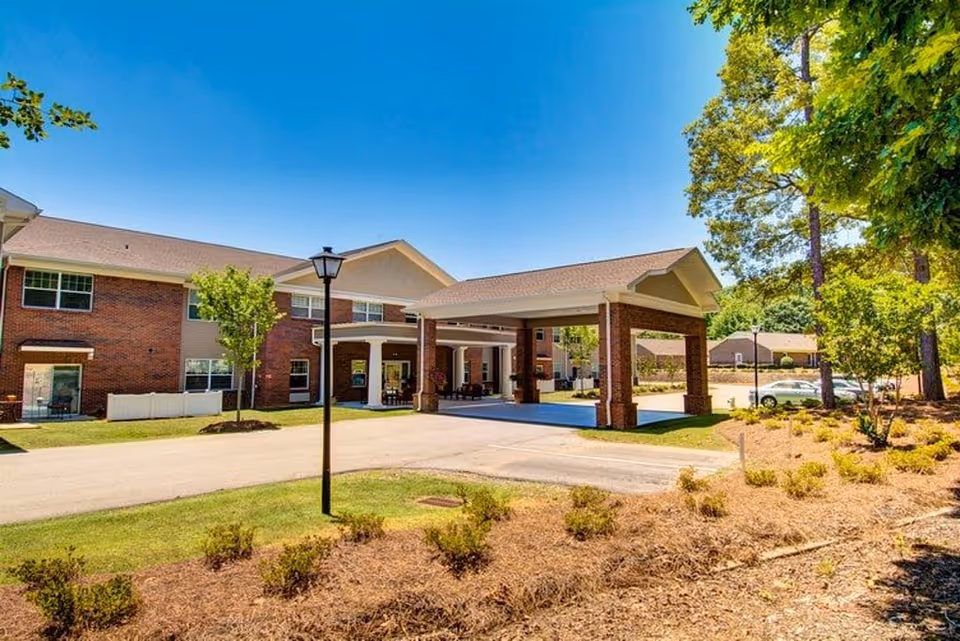 Exterior view of a senior living facility building with a covered entrance driveway, surrounded by trees and landscaping under a clear blue sky.