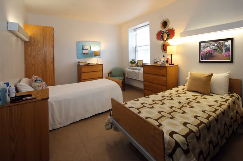 A cozy bedroom in a senior living facility with two single beds, each with wooden headboards and footboards. One bed has a white bedspread and a decorative pillow, while the other has a patterned bedspread with a beige pillow. The room features two wooden dressers, a green armchair near a window with blinds, and various decorative items including framed artwork, a lamp, and wall-mounted masks. The walls are painted white, and the floor is covered with a light brown linoleum.