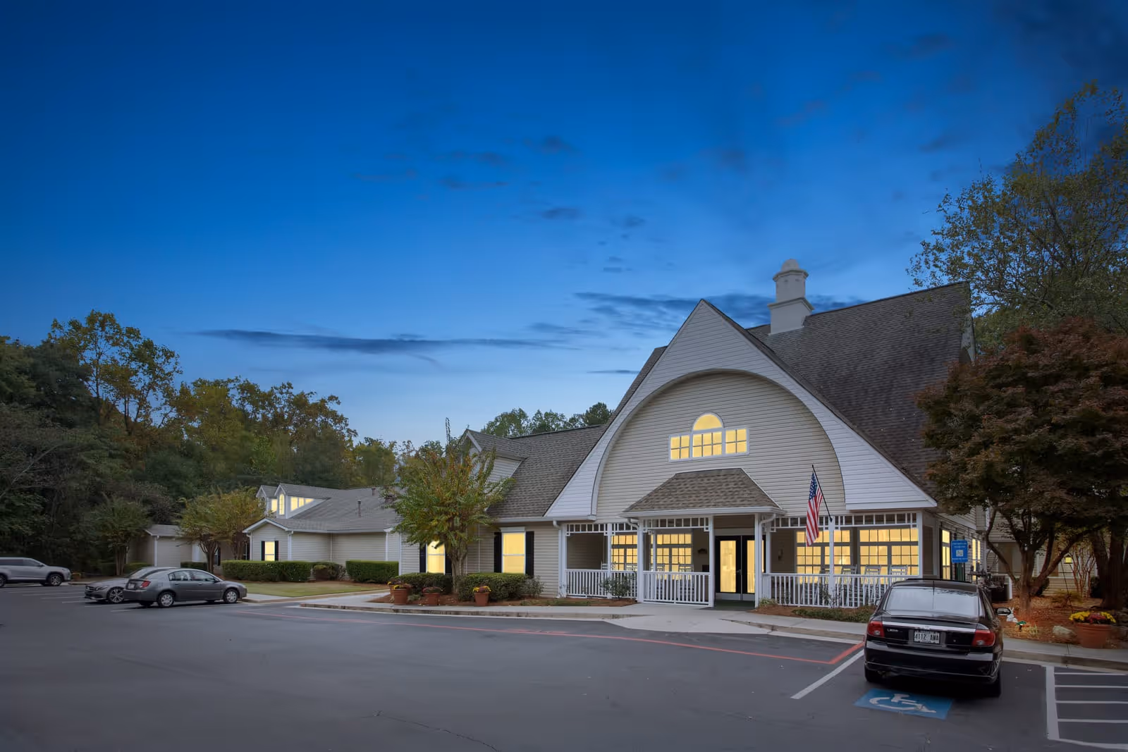 Exterior view of Governor's Glen Memory Care & Assisted Living Community building at dusk with lights on inside. The building has a large peaked roof, white siding, and a front porch with an American flag. Several cars are parked in the parking lot in front, including one in a handicapped parking space. Trees and greenery surround the facility.