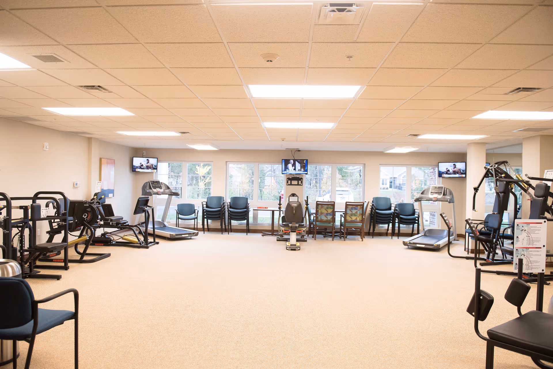 A spacious fitness room with various exercise equipment including treadmills, weight machines, and stationary bikes. Several chairs are stacked and arranged along the windows, and three televisions are mounted on the walls. The room has large windows letting in natural light and a beige carpeted floor.