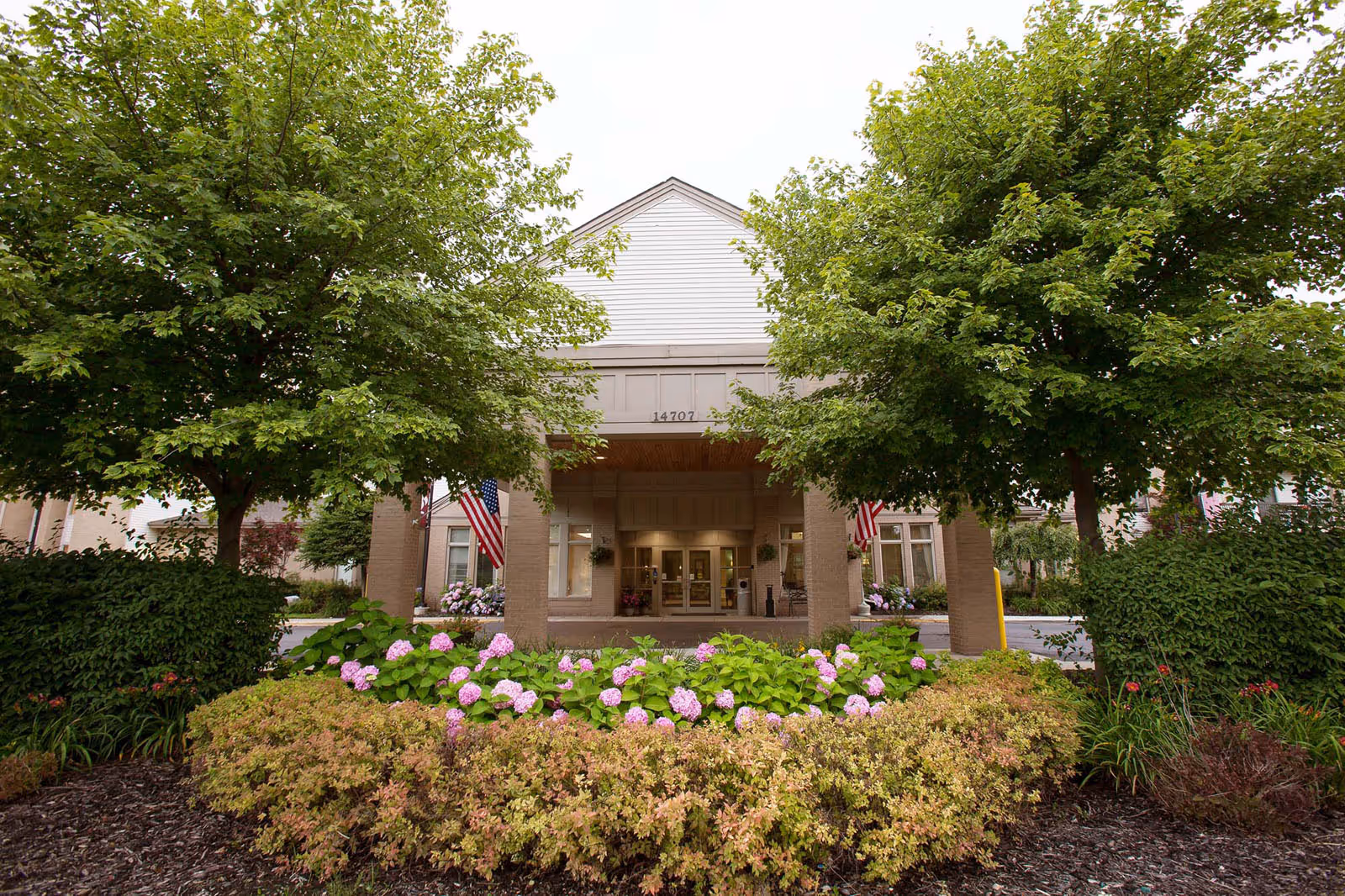 Front entrance of a senior living building with a covered portico, American flags, and landscaped flower beds.