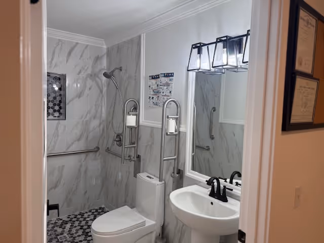 Marble-tiled bathroom with toilet, sink, grab bars, and shower in a senior living facility.