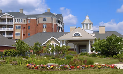 Exterior view of Mercy Ridge facility showing a multi-story brick and beige building with a covered entrance, surrounded by green lawns, colorful flower beds, and trees under a partly cloudy blue sky.