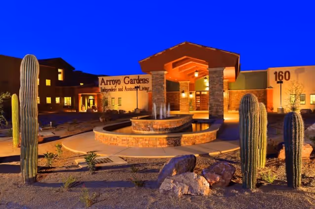 Exterior view of Arroyo Gardens Independent and Assisted Living facility at dusk, featuring a stone fountain in front, desert landscaping with tall cacti and rocks, and the building entrance illuminated with warm lighting.