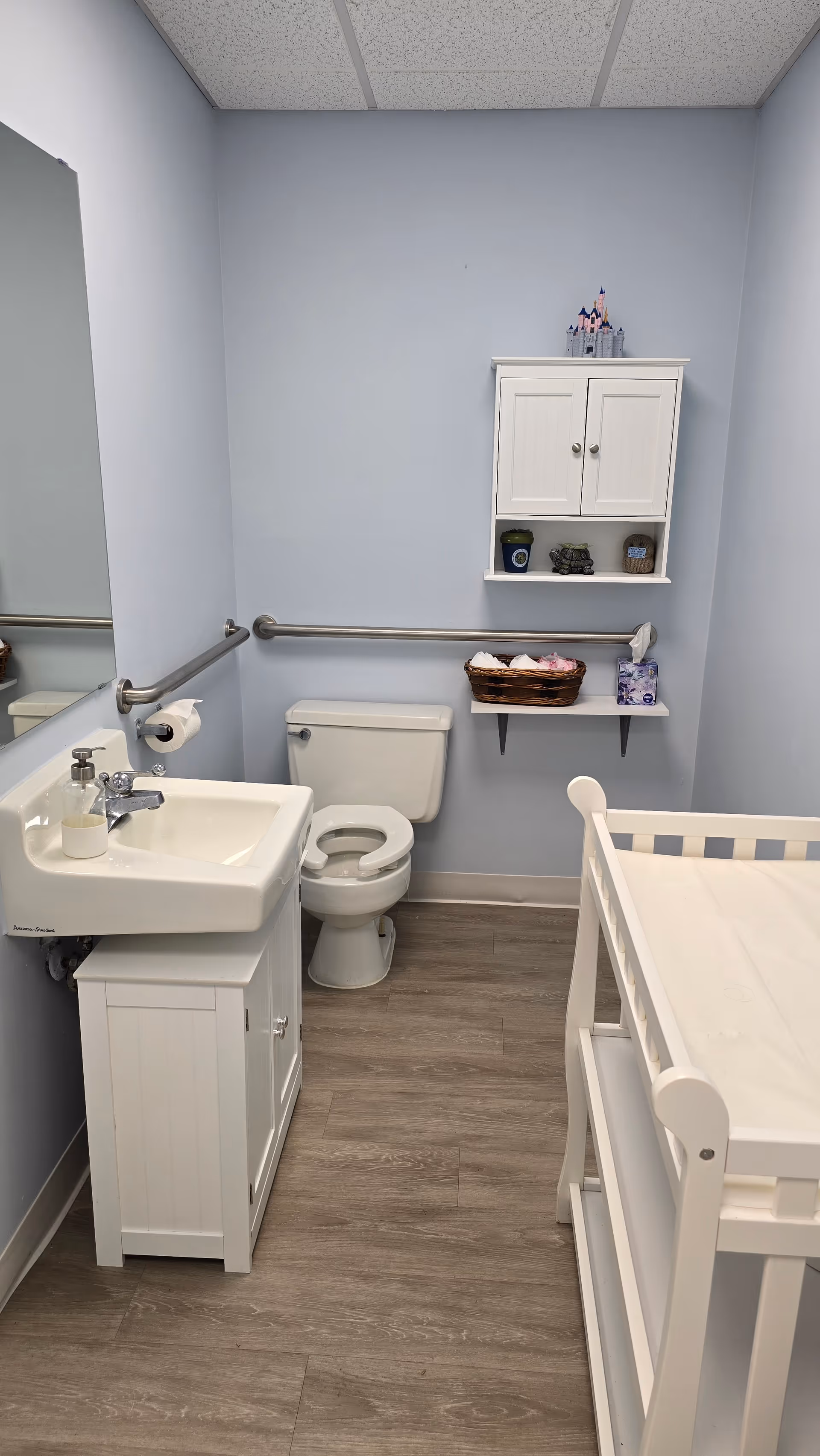 A small light-blue bathroom with sink, toilet, shelf, and white crib.