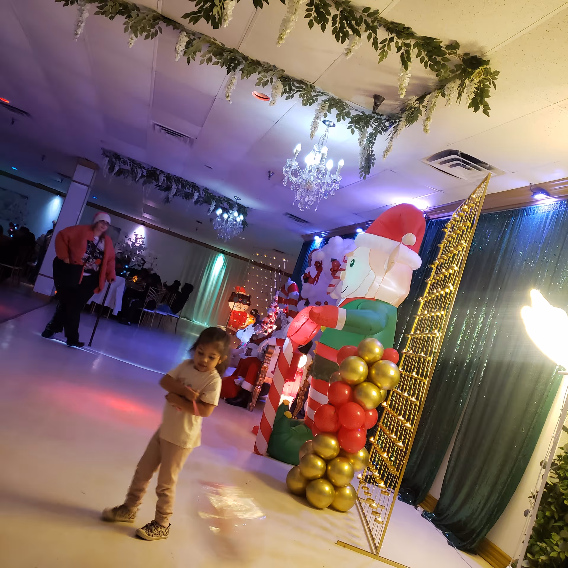 Indoor party hall with holiday decorations, balloons, inflatables, and a child in foreground.