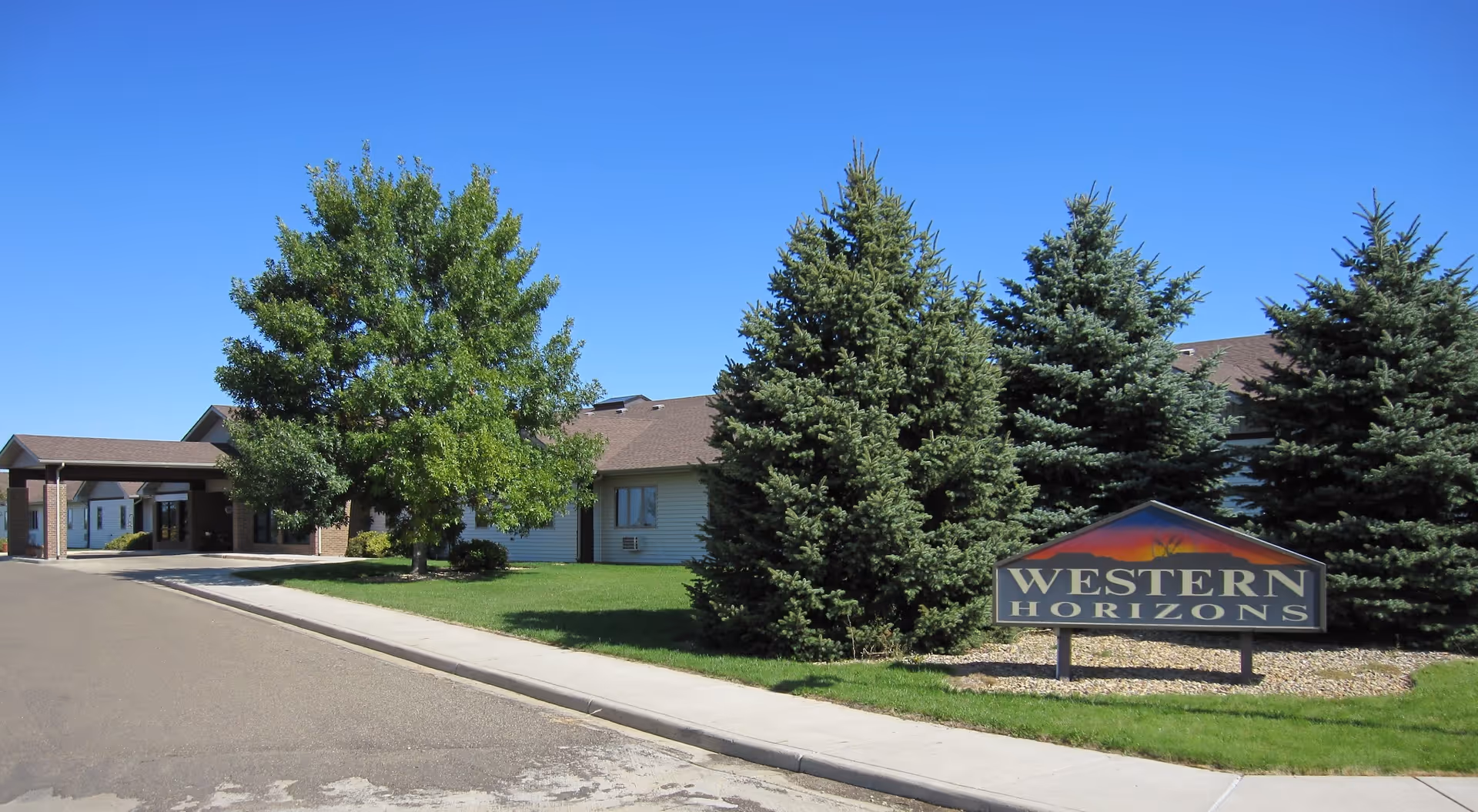 Exterior view of Western Horizons Assisted Living facility with a driveway, green lawn, several trees, and a sign displaying the facility's name under a clear blue sky.