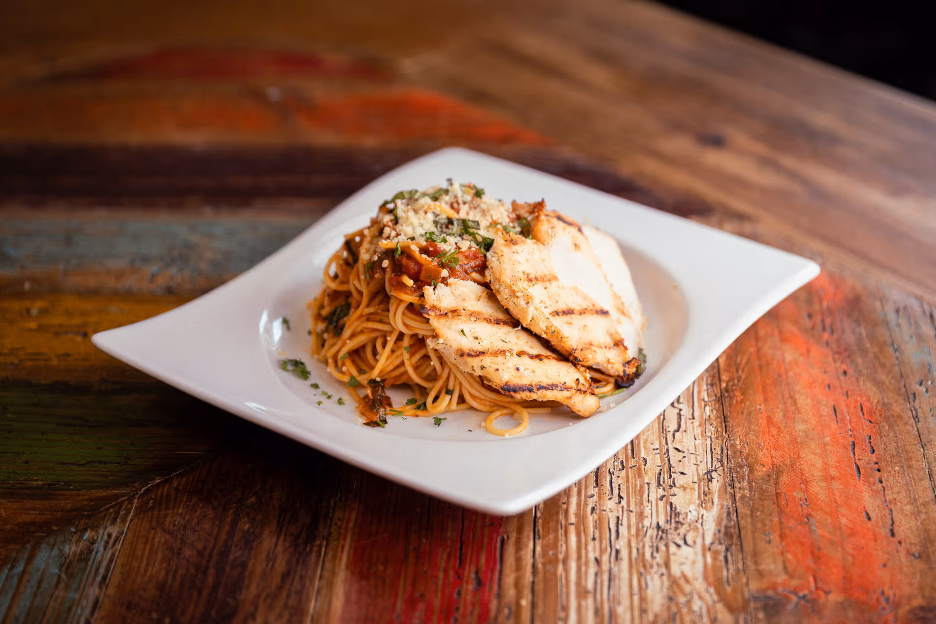 A white square plate with grilled chicken breast slices served on a bed of spaghetti pasta topped with tomato sauce, grated cheese, and herbs on a rustic wooden table.