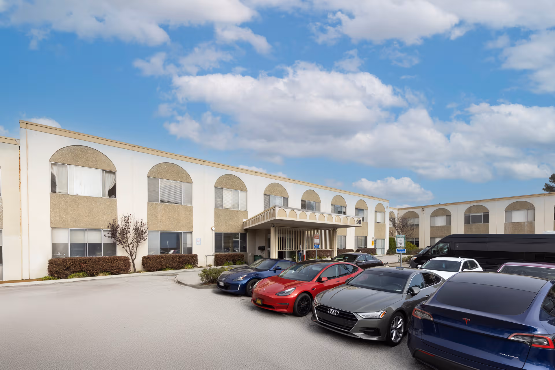 Beige, two-story senior living facility with arched windows, cars parked, and a blue sky.