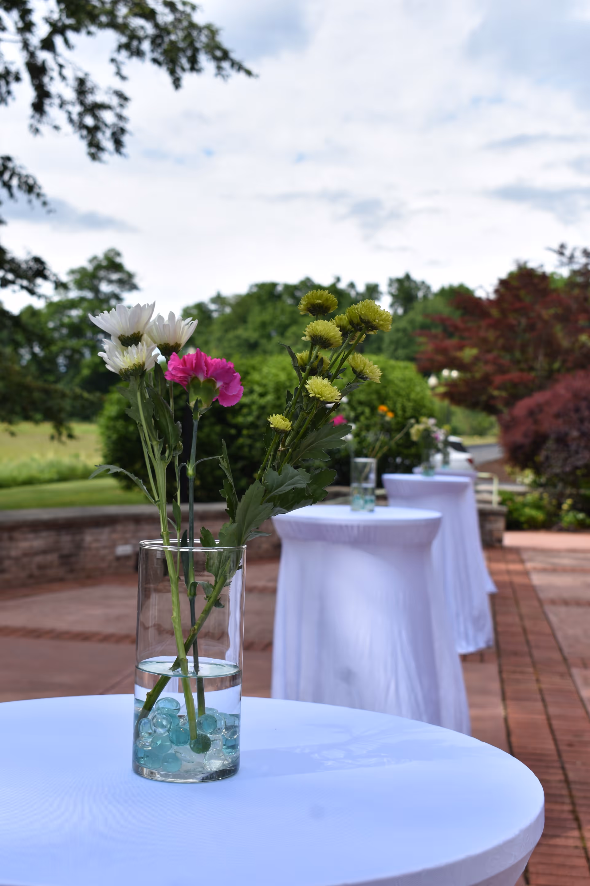 Patio with tall white-clothed cocktail tables topped with glass vases of flowers against a garden backdrop.