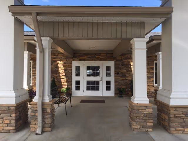 Entrance to a building with a covered porch supported by white columns with stone bases. The entrance has white double doors with glass panels and a brown doormat in front. There is a black metal bench to the left side of the entrance.