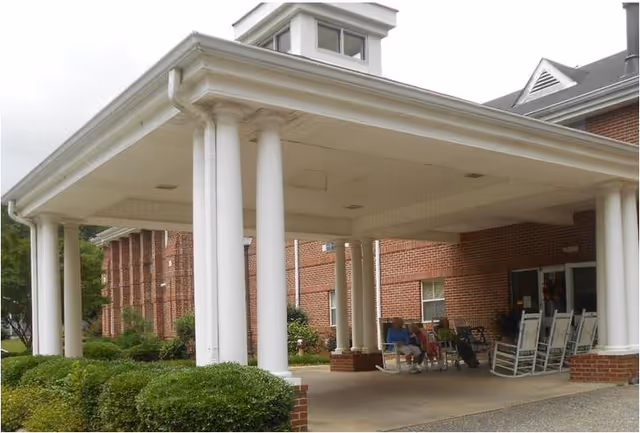 Covered entrance area of a brick building with white columns and a small cupola on the roof. There are bushes along the side and several rocking chairs under the covered area, with two people sitting in the chairs near the entrance.