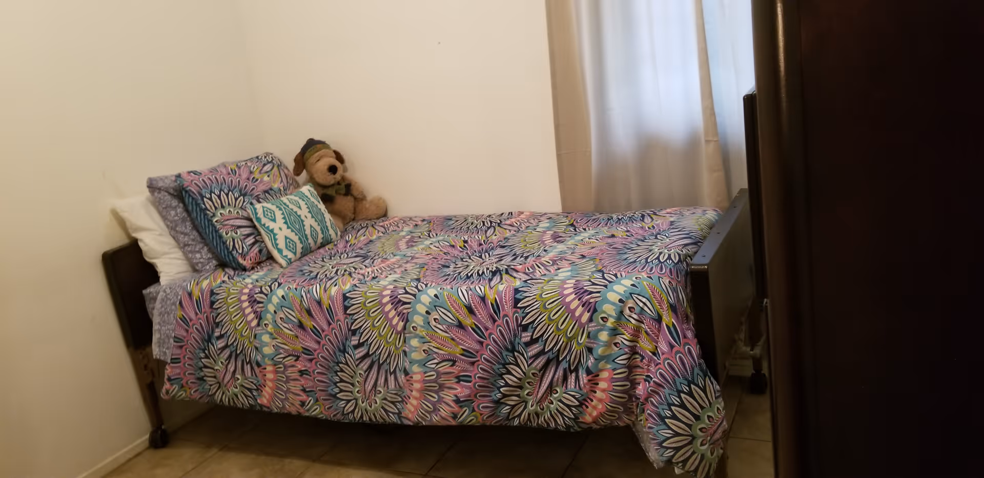 A small bedroom with a single bed, colorful quilt, pillows, and a teddy bear.