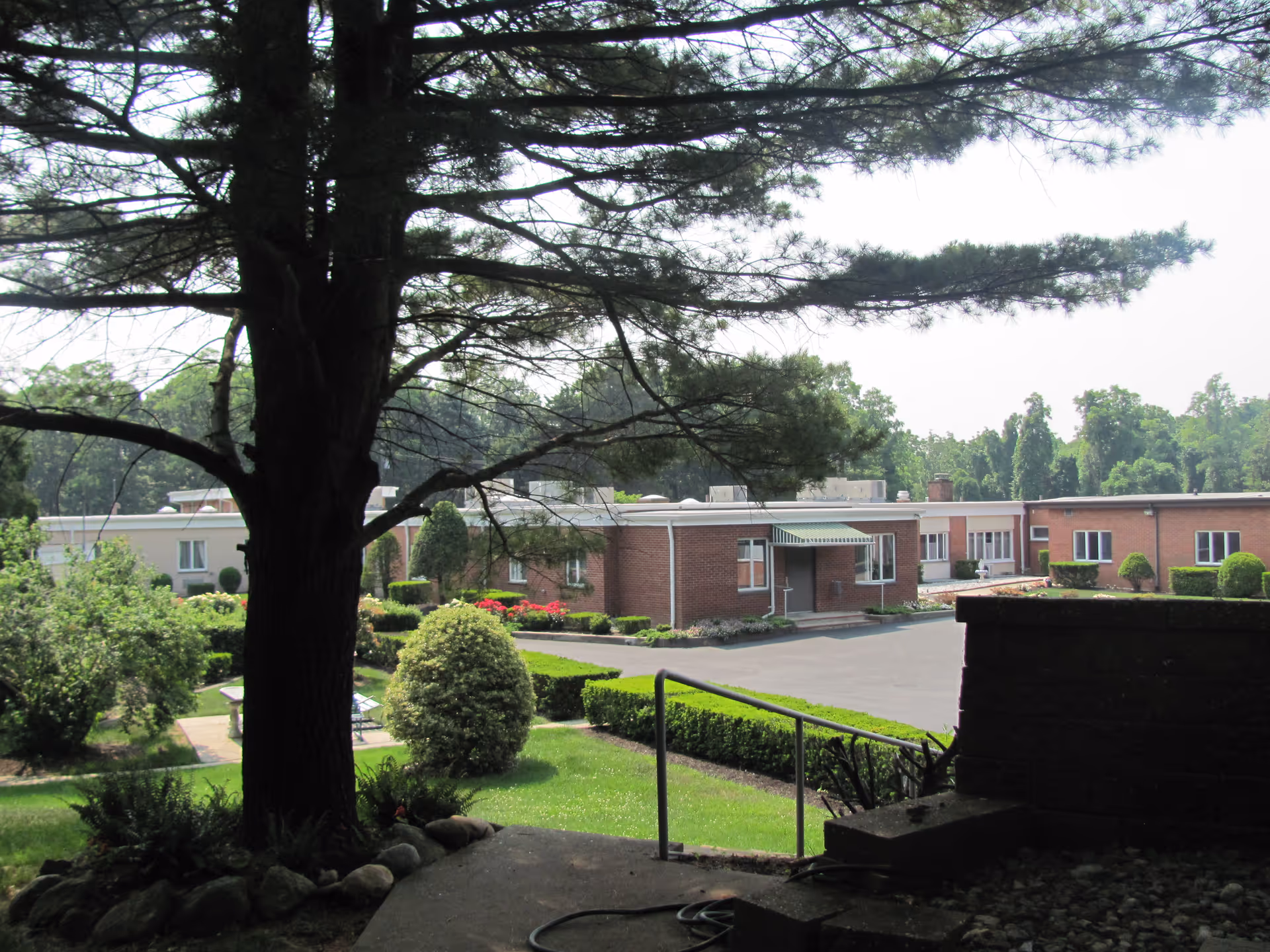 View of a single-story brick building and landscaped courtyard framed by a large tree in the foreground.