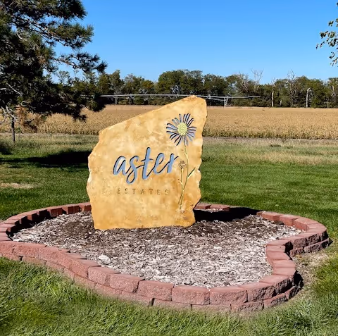 A large stone sign reading 'Aster Estates' sits in a circular mulch bed on manicured lawn.