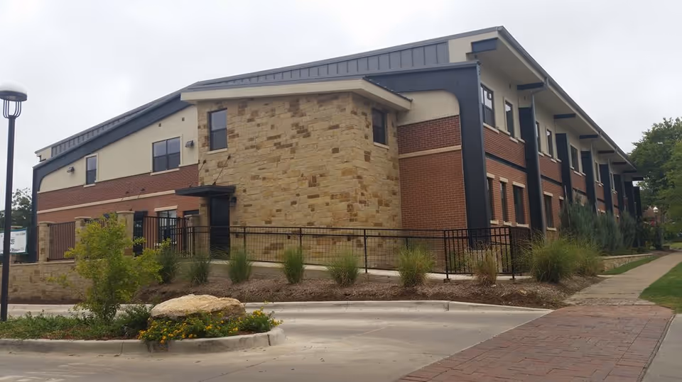 Exterior view of a two-story senior living facility building with a combination of brick and stone facade, black framed windows, and a sloped roof. The building is surrounded by landscaped greenery, a sidewalk, and a paved driveway under an overcast sky.