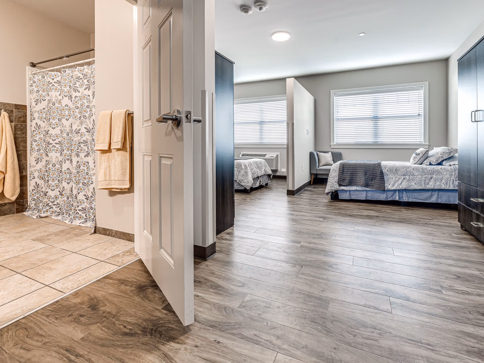 View of a senior living facility room showing two beds separated by a partial wall, a window with blinds, a chair, and a bathroom with a shower curtain and towels visible through an open door. The room has wood-style flooring and neutral-colored walls.