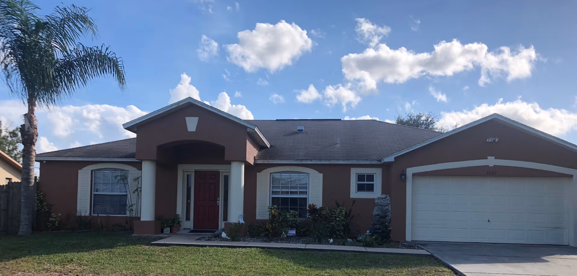 Single-story tan assisted living facility with red entrance door, palm tree, and two-car garage.