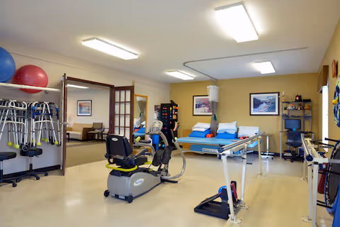 Rehabilitation gym inside a senior living facility with exercise equipment and a bed.