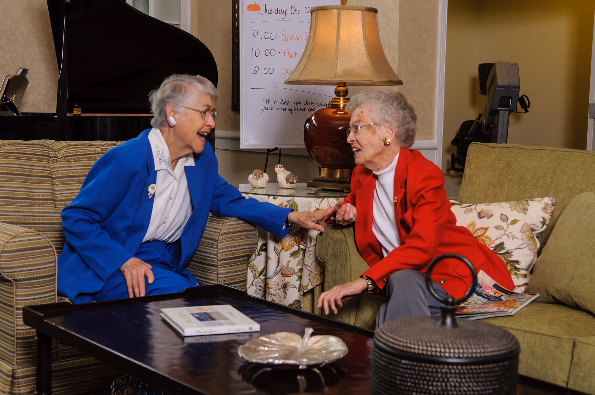 Two elderly women sitting and chatting in a cozy living room area. One woman is wearing a blue suit and the other a red jacket. They are smiling and holding hands across a coffee table with a book and decorative items. Behind them is a lamp, a piano, and a whiteboard with a schedule written on it.