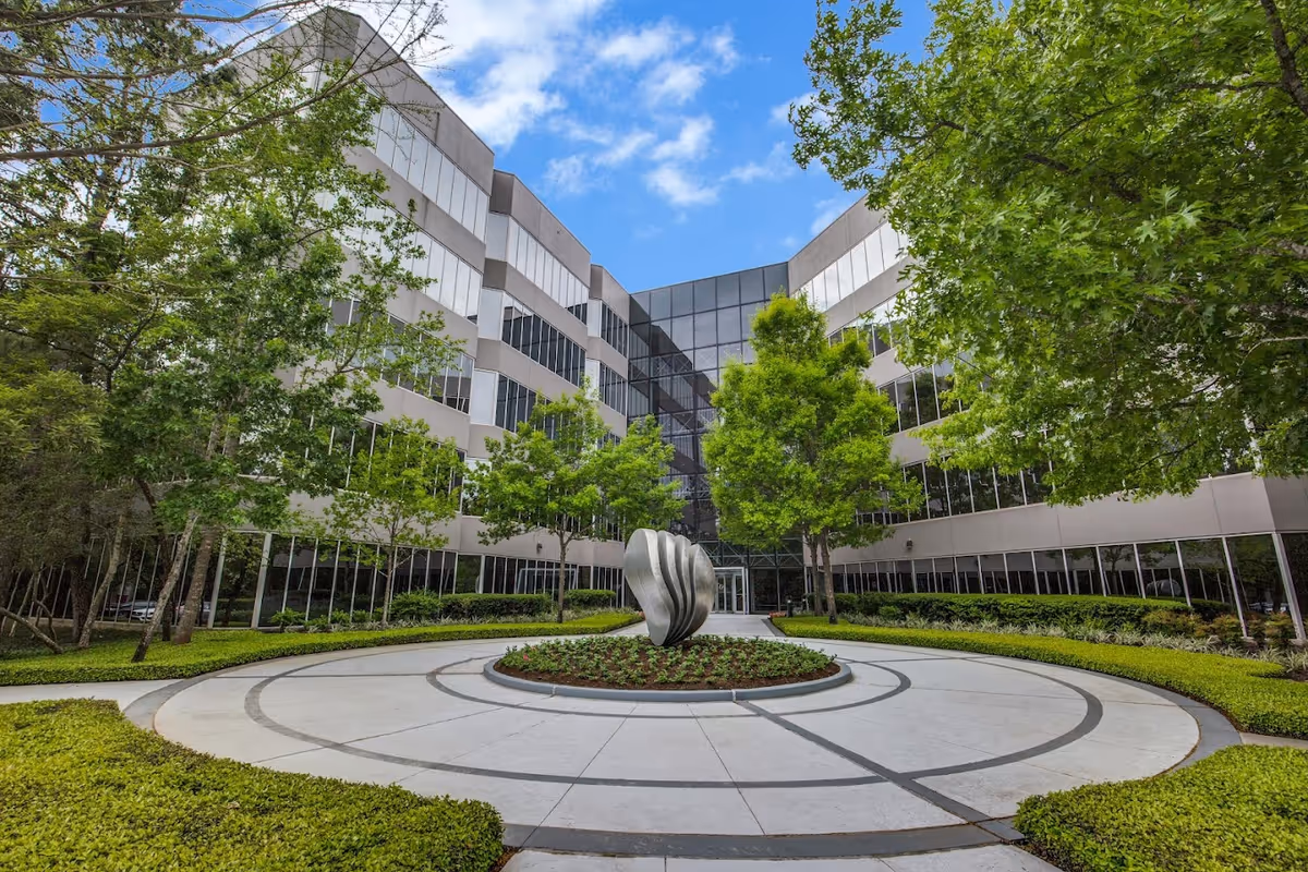 Modern exterior courtyard with a circular tiled plaza, sculpture centerpiece, and surrounding glass-faced building.