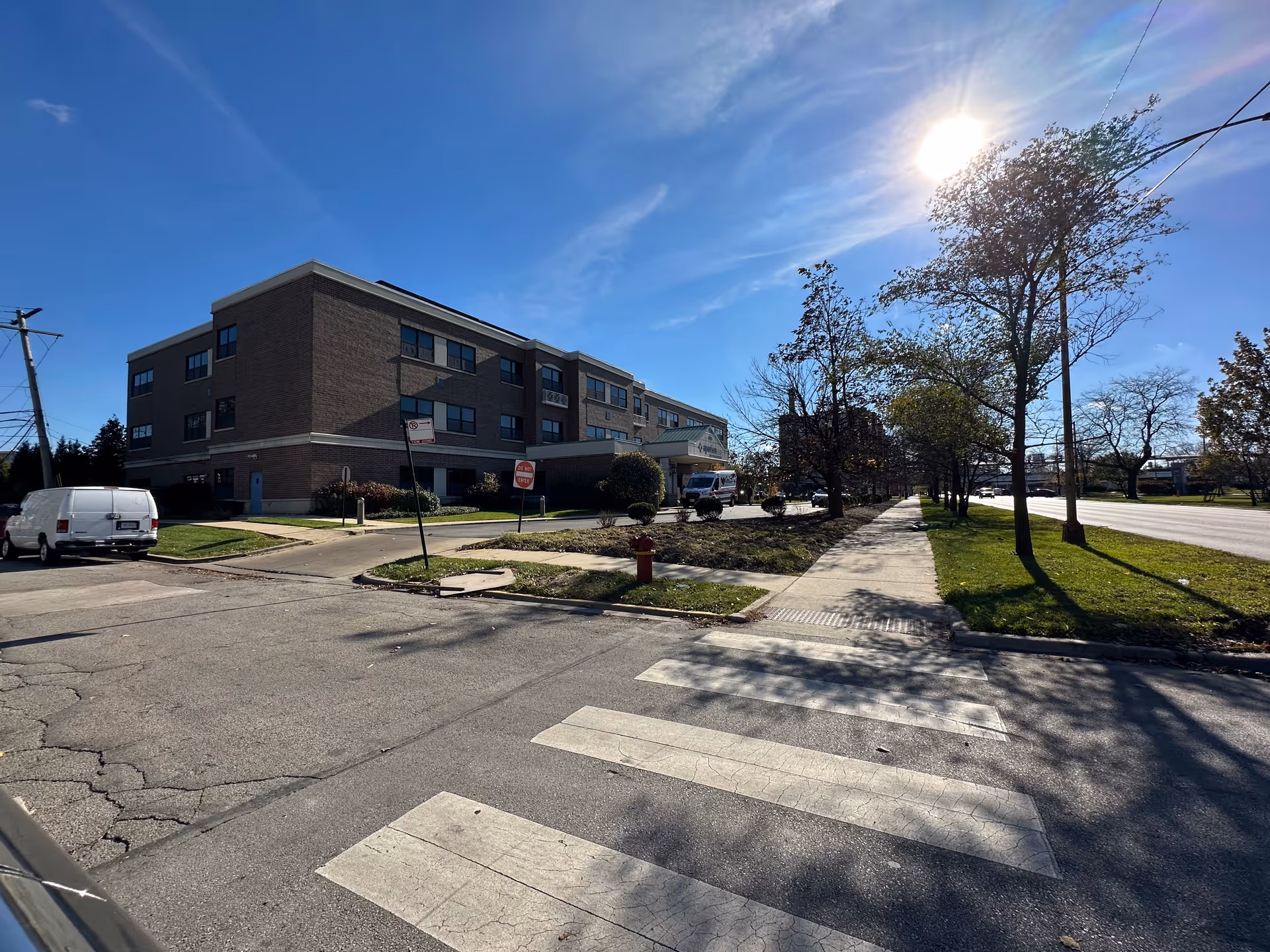 Exterior view of a three-story brick building under a clear blue sky with the sun shining brightly. The building has multiple windows and a covered entrance with a sign that reads Aperion Care International. There is a sidewalk, a crosswalk, some trees, and a white van parked near the building.