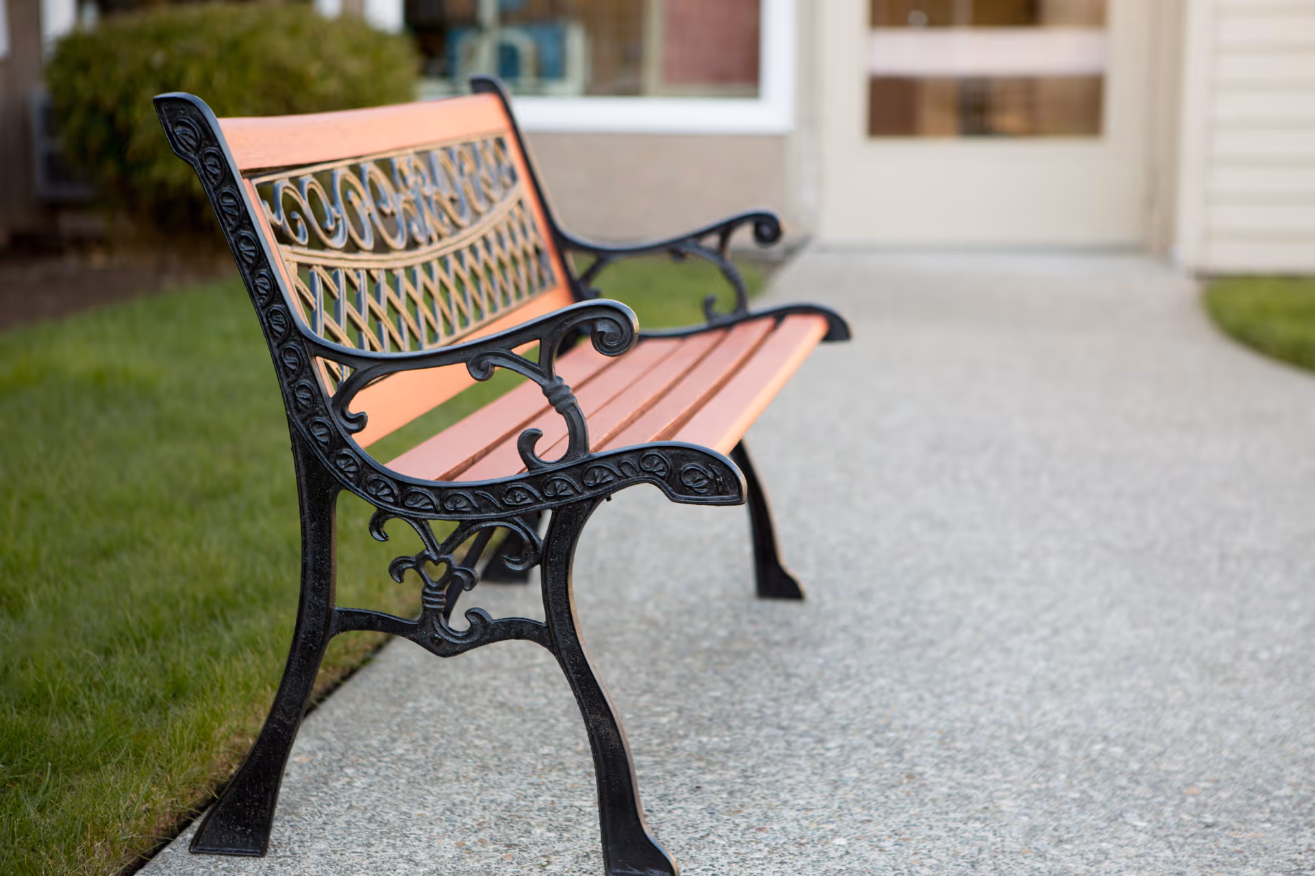 A decorative outdoor bench with wooden slats and ornate black metal armrests and legs, placed on a concrete pathway next to a grassy area in front of a building entrance.