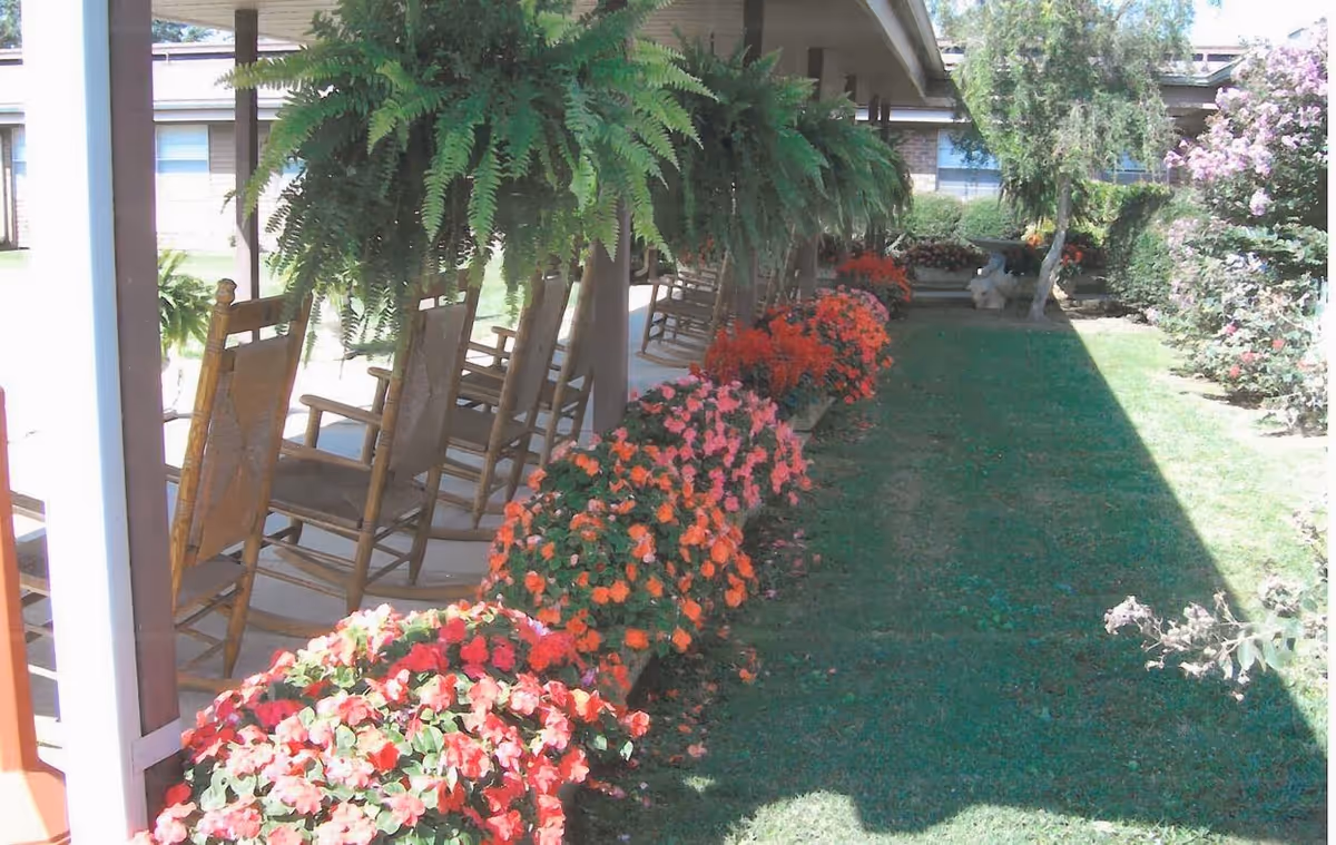 Covered porch with wooden rocking chairs, hanging ferns, and a row of colorful flowers along a grassy lawn.