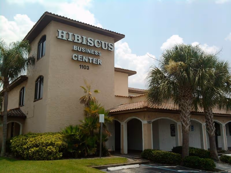 Beige two-story building with Hibiscus Business Center sign, arches, and palm trees in front.