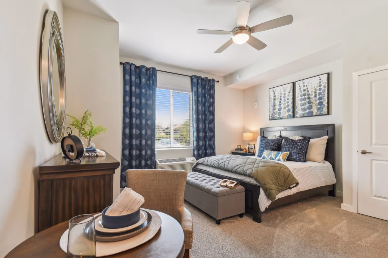 Well-lit furnished bedroom with a bed, decorative pillows, an upholstered bench, a dresser with mirror, a small table and chair, and a window with blue curtains.