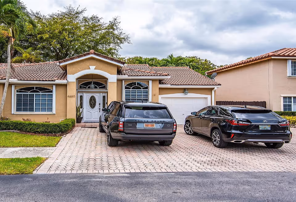 Front exterior view of a single-story house with a tiled roof, two large windows, a white front door with glass panels, and a driveway with two black cars parked on it. There is greenery including bushes and trees around the house.