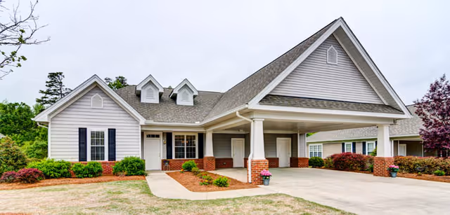 Front exterior of a single-story senior living building with a covered porte-cochere entrance, driveway, and landscaped lawn.