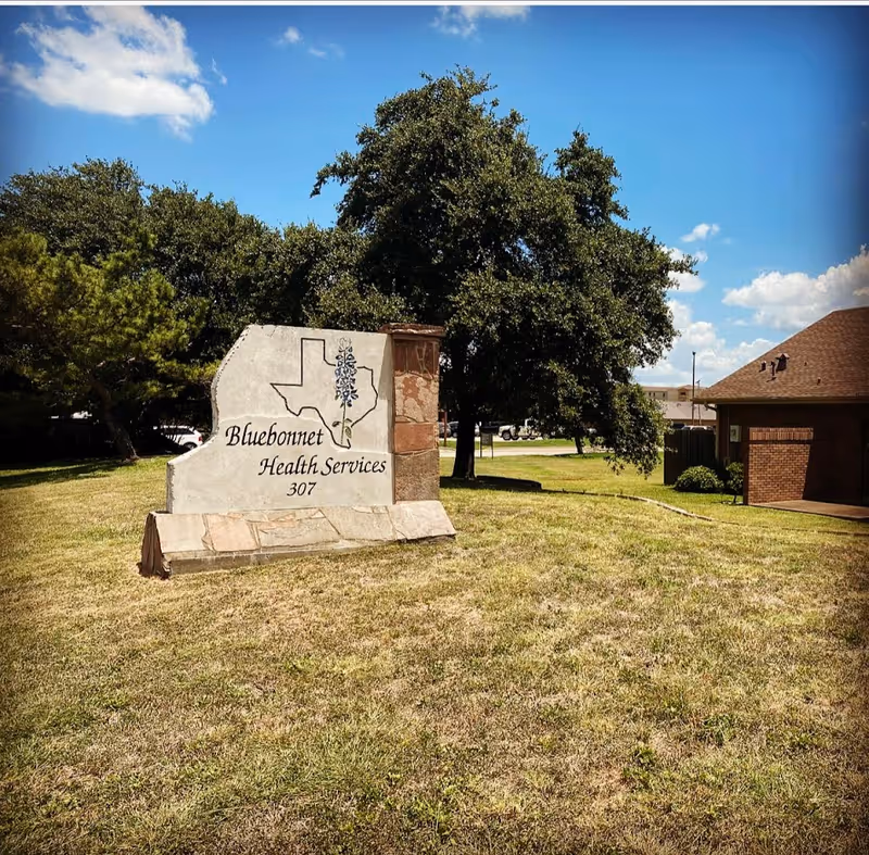 A stone sign reading Bluebonnet Health Services Hospice sits on a grassy lot beside a building