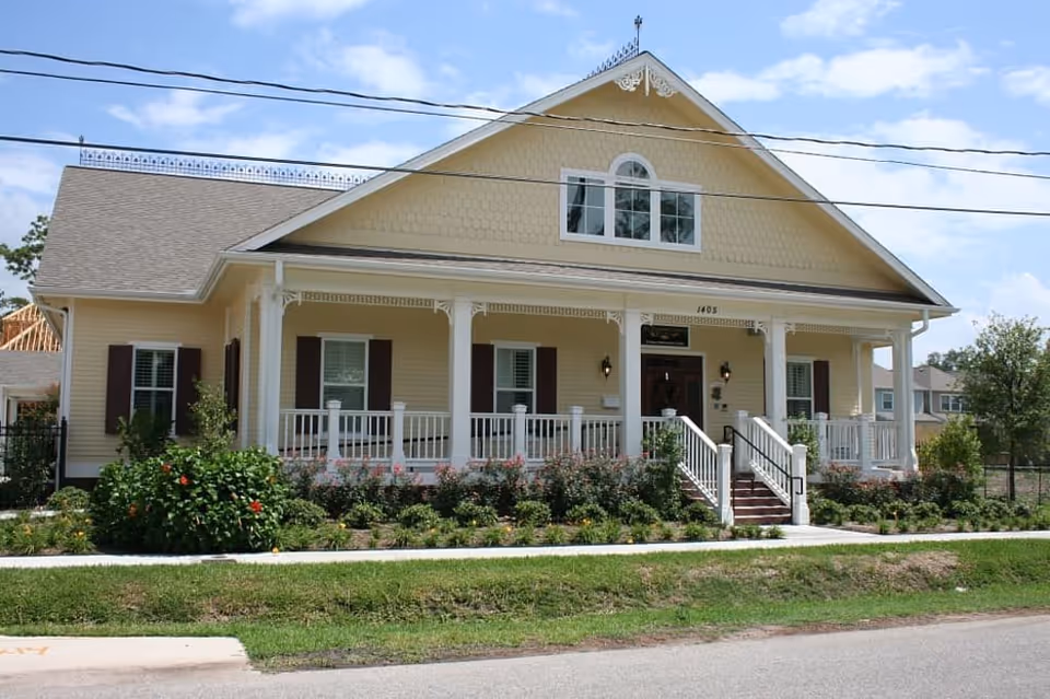 Front exterior view of a yellow single-story house with a large porch, white railings, and steps leading up to the entrance. The house has multiple windows with brown shutters and is surrounded by a well-maintained garden with bushes and flowers. The sky is partly cloudy.