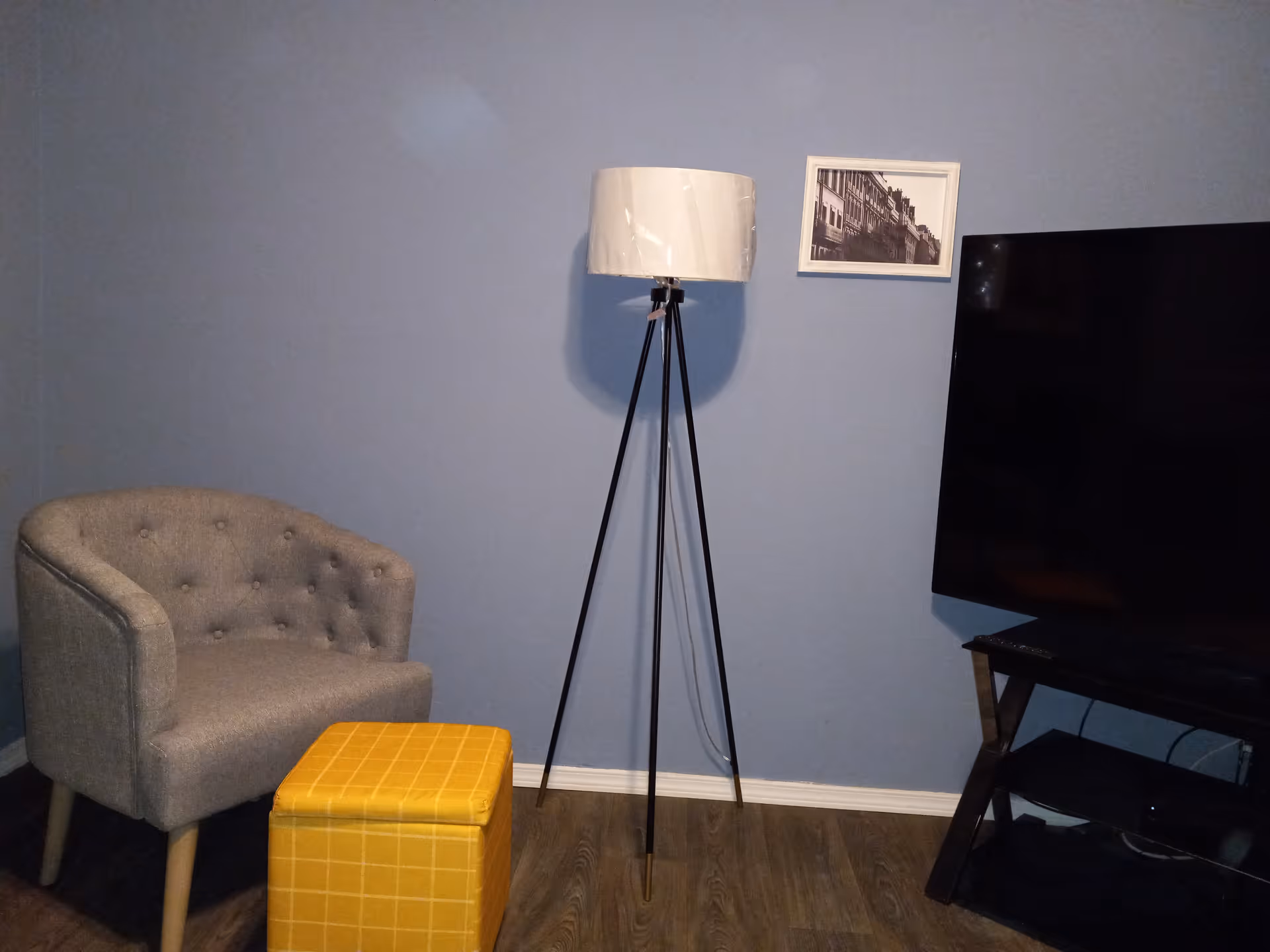 A blue-walled living room corner with chair, yellow ottoman, tripod lamp, and TV.