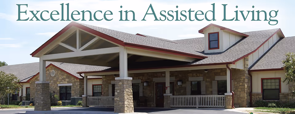Front entrance of an assisted living building with a covered porte-cochere and stone facade beneath the words "Excellence in Assisted Living".