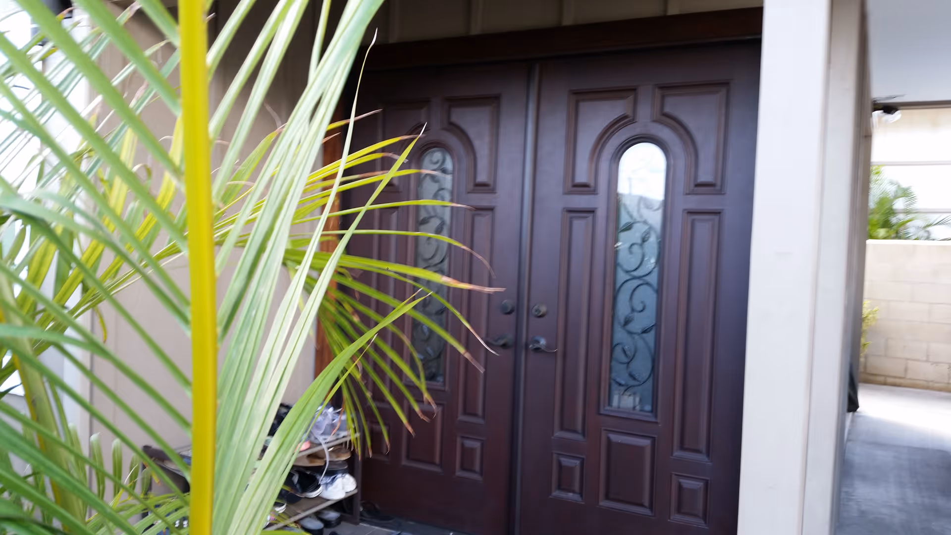 Double wooden front doors with decorative glass panels, partially obscured by green palm leaves in the foreground. The entrance is part of a building with beige walls and a covered walkway visible to the right.
