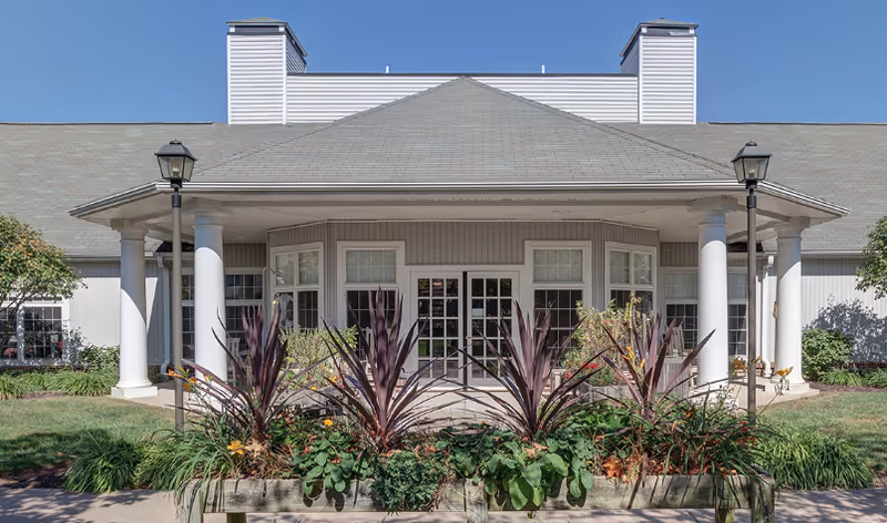 Front entrance of a light-colored community building with a covered porch supported by white columns, glass double doors, and a planted flowerbed in front.
