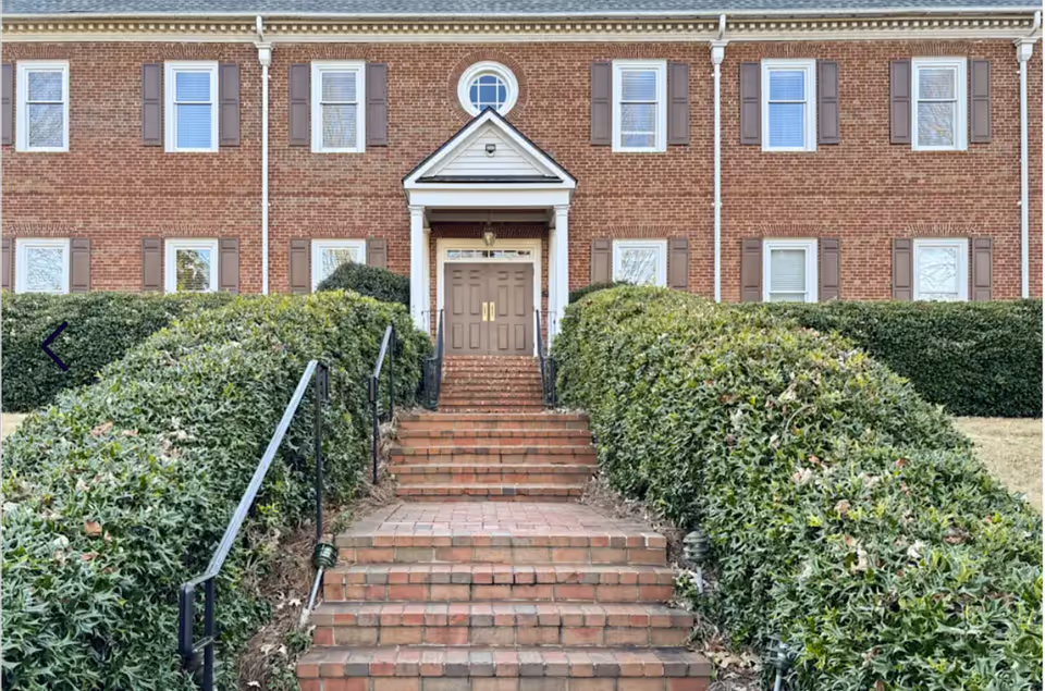 Brick building front with brick stairs, railing, and neatly trimmed hedges leading to a double door.