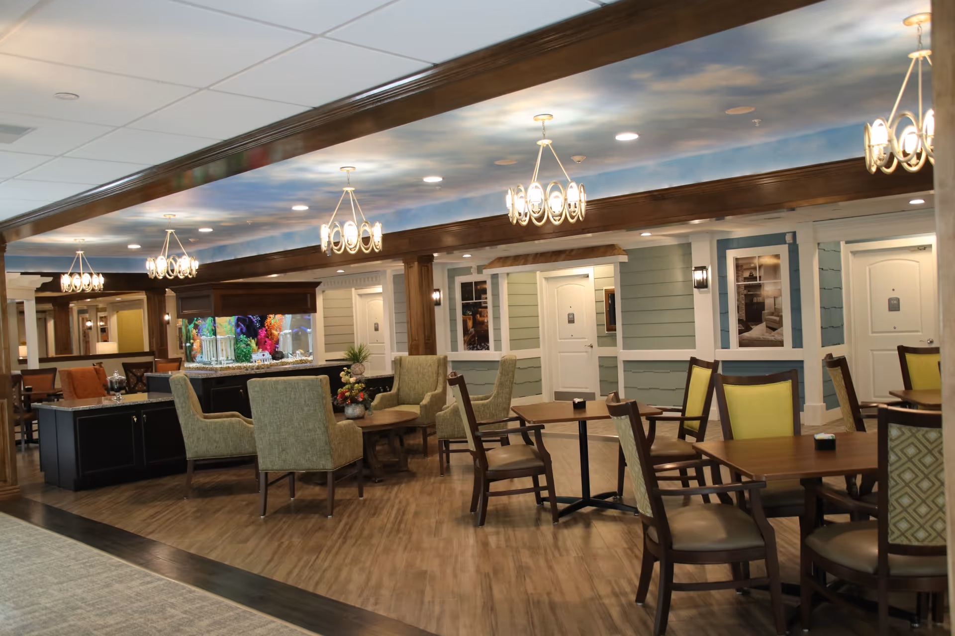 A spacious and well-lit common area in a senior living facility featuring multiple seating arrangements with wooden tables and cushioned chairs. The room has a decorative ceiling with chandeliers, wood beams, and a large aquarium as a centerpiece. The walls have a light green and white panel design with framed pictures and several white doors.