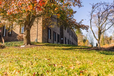 A brick senior living facility sits beside a grassy lawn with autumn trees.