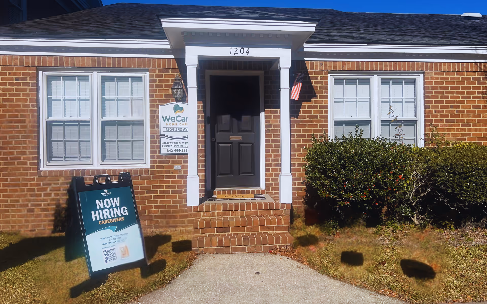 Brick front of WeCare Home Care building with entrance, two windows, and a 'Now Hiring' sign.