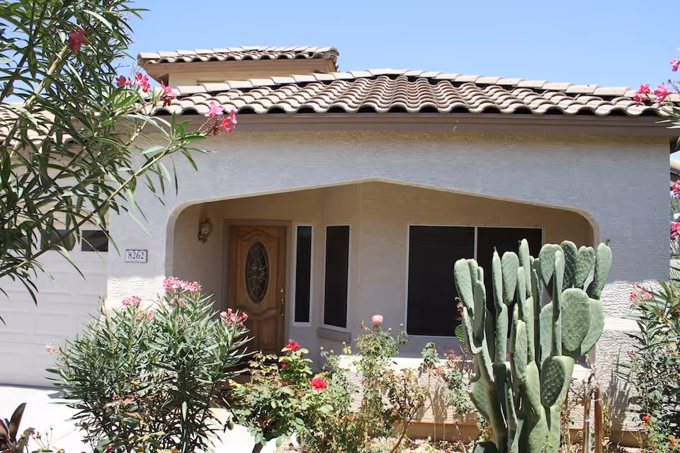Front entrance of a stucco house with a tiled roof, wooden door, large cactus and flowering shrubs in the yard.