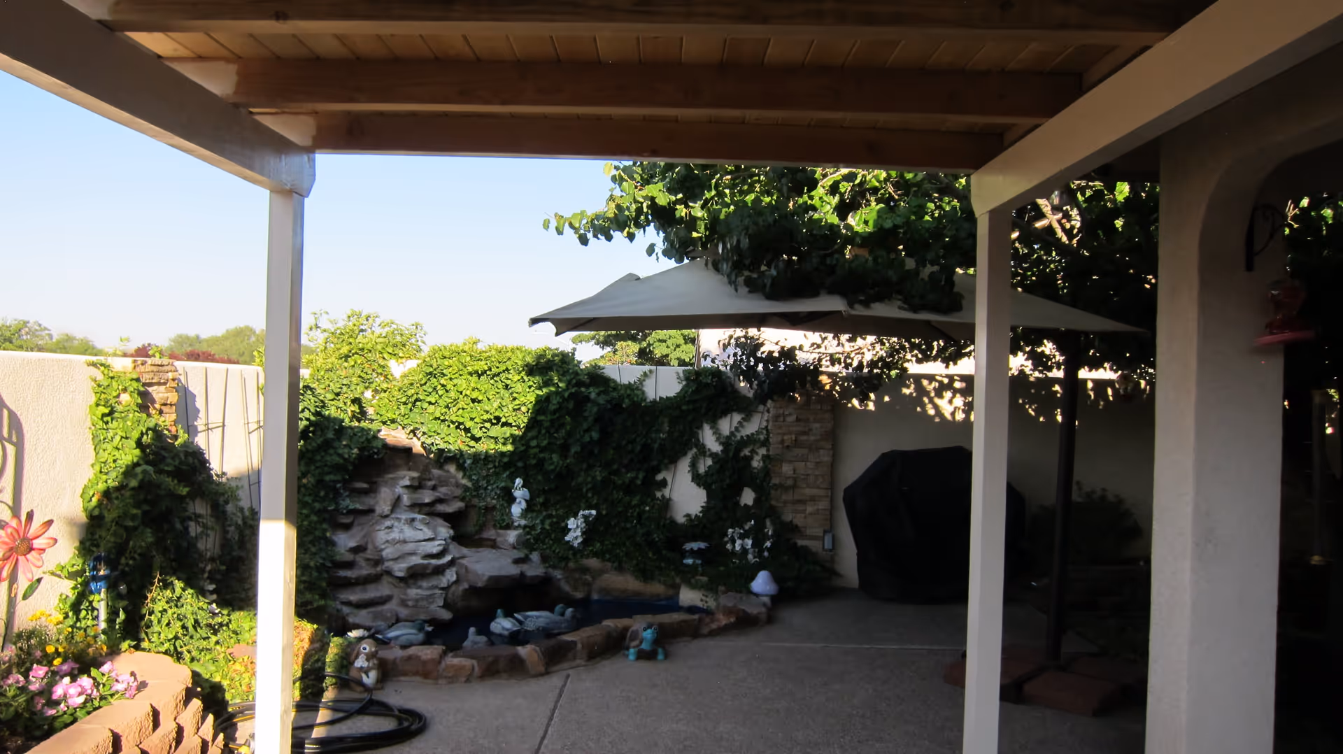 Covered patio area with wooden ceiling and white support beams overlooking a small garden pond with decorative rocks, greenery, and garden ornaments. There is a large umbrella and a covered grill in the background.